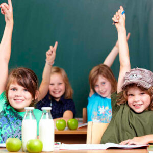 Kinder an Tischen sitzend mit Milchflaschen und Äpfel daraufstehend strecken einen Arm in die Höhe um sich zu melden